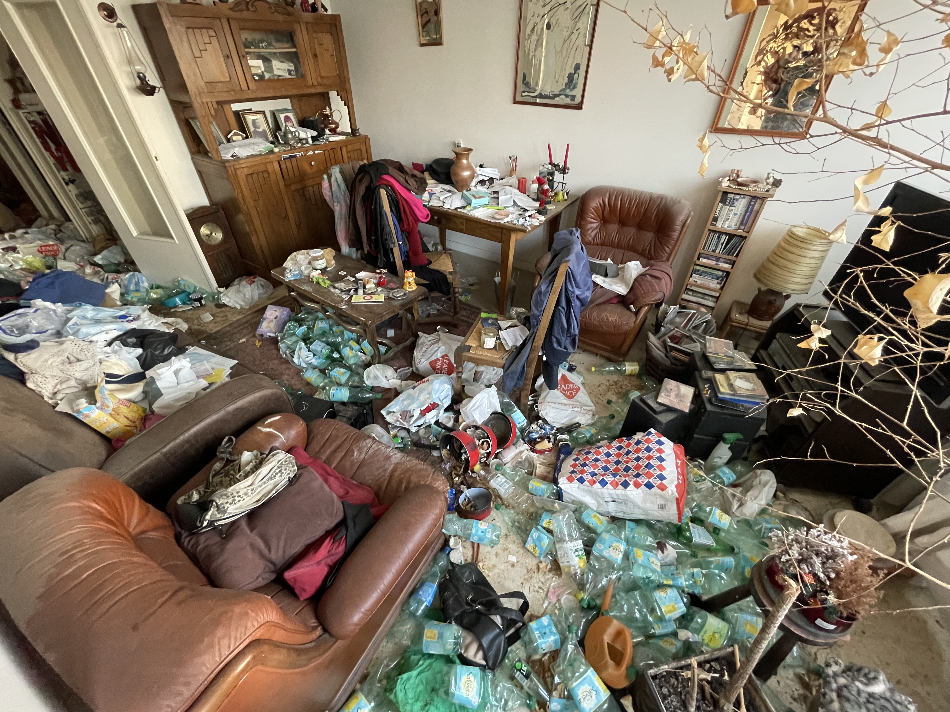 Salon en syndrome de Diogène : accumulation de centaines de bouteilles plastique, fauteuils en cuir, vaisselier ancien — appartement encore meublé