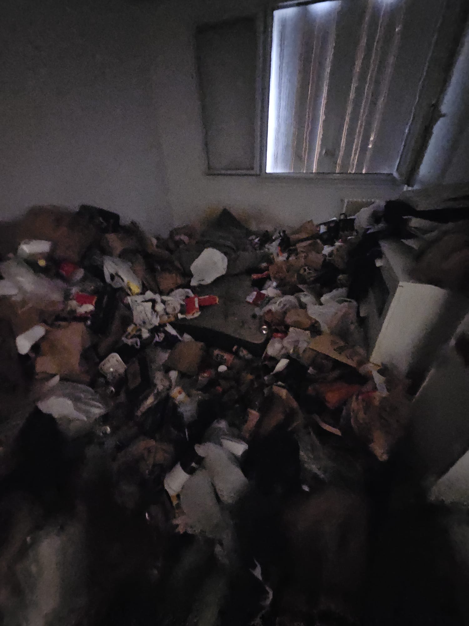 Pièce sombre avec accumulation hétéroclite de cartons et déchets, peu de lumière naturelle